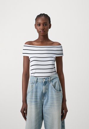 Off-the-shoulder witte top met zwarte horizontale strepen. Getextureerde geribbelde stof, gecombineerd met lichtblauwe, loszittende denim jeans.