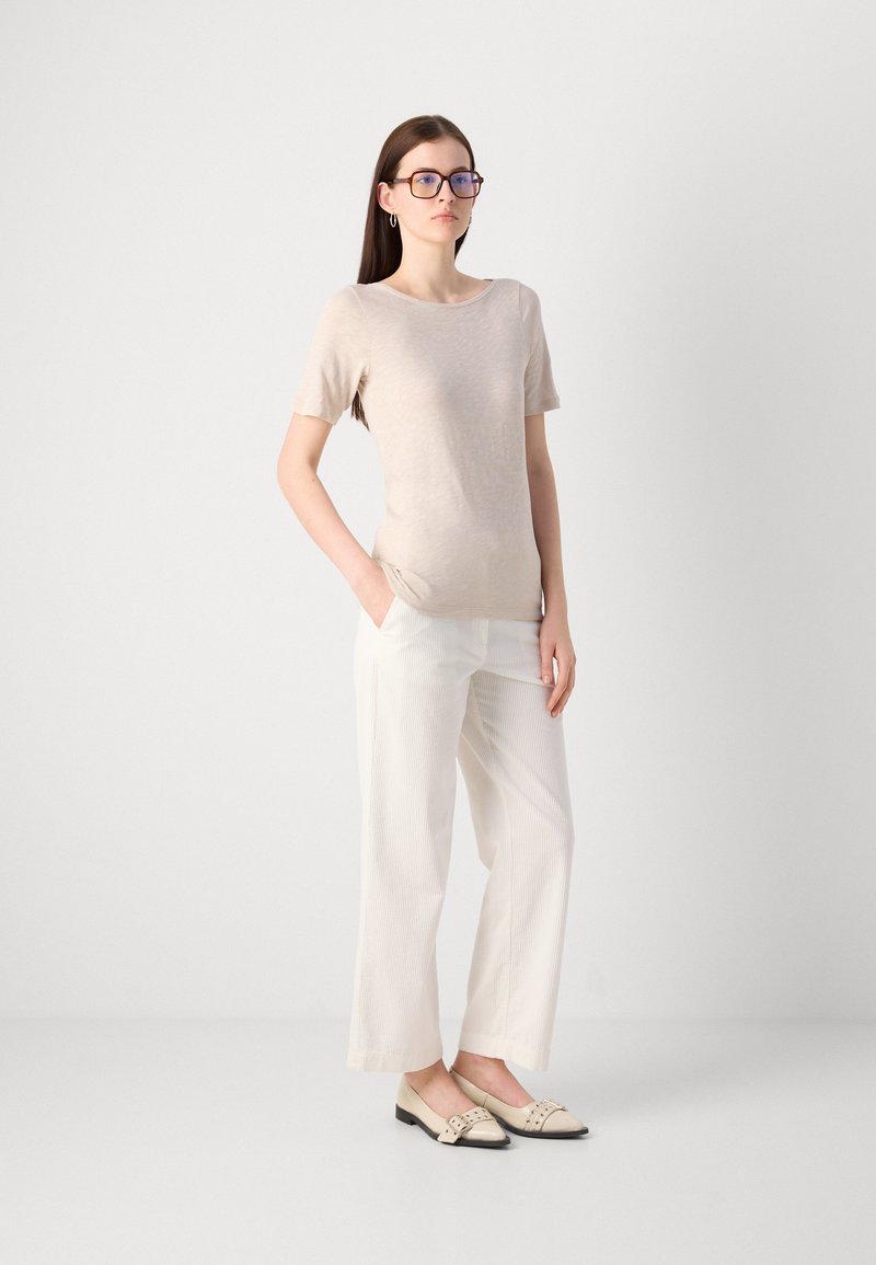 Beige Kurzarmoberteil, hellgestreifte weite Hose und cremefarbene Ballerinas mit schwarzen Akzenten. Das Model posiert mit einer Hand in der Tasche.