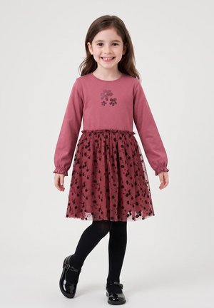 Junges Mädchen lächelt, trägt ein langärmeliges pinkes Kleid mit floralem Rock, schwarze Strumpfhosen und schwarze Mary Jane Schuhe, steht vor einem einfarbigen Hintergrund.
