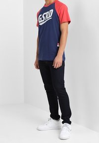 Námořnická modrá a červená polo tričko s krátkým rukávem s logem "GSRD", kombinované s černými džíny a bílými teniskami. Uvolněný střih a bavlněný materiál.