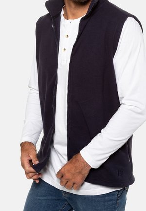 Mężczyzna w białej koszuli z długim rękawem pod czarną kamizelką na zamek błyskawiczny i niebieskimi dżinsami, poprawia zamek kamizelki na wysokości talii.