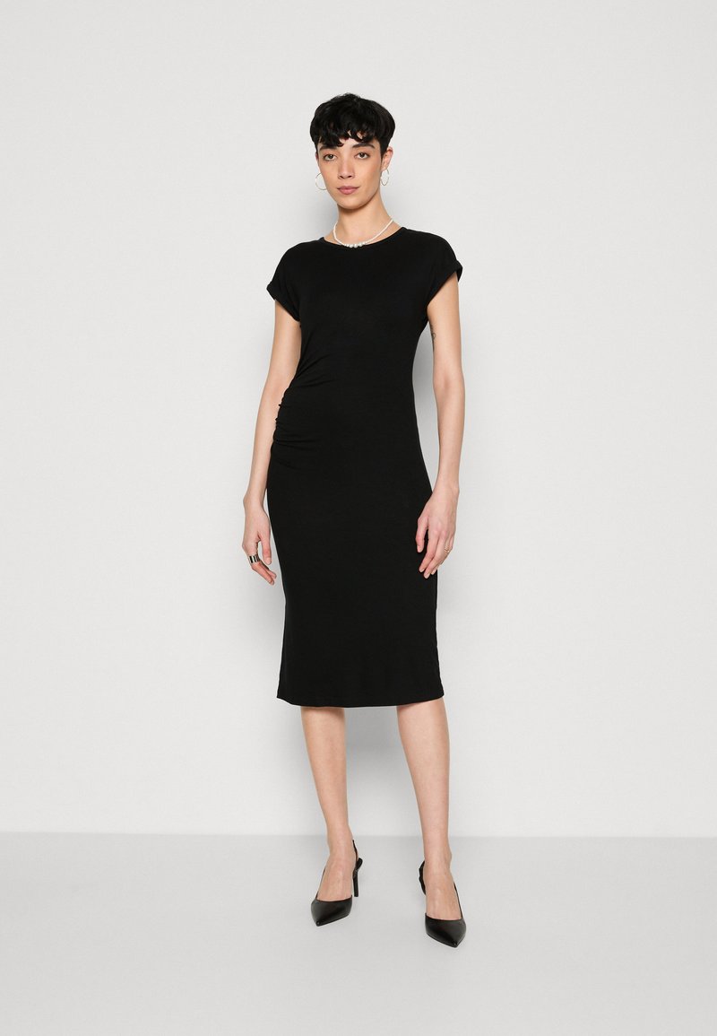Czarna, dopasowana sukienka z krótkimi rękawami, do kolan, wykonana z gładkiej tkaniny, z delikatnym marszczeniem z boku, zestawiona z czarnymi szpilkami.