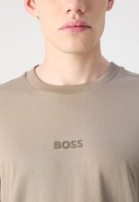 Szürke, rövid ujjú póló, puha anyagból készült, bordázott kerek nyakkivágással és dombornyomott "BOSS" logóval a mellkasán.