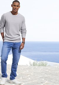 Hellgrauer strukturierter Pullover, blaue Jeans und weiße Sneaker. Mann steht auf einem steinigen Untergrund mit dem Ozean im Hintergrund, die Hände entspannt an den Seiten.