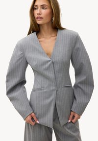 Femme portant un costume gris à fines rayures avec des manches surdimensionnées, serrant le devant de la veste et regardant légèrement sur le côté.