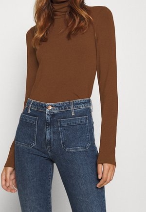Kvinde iført brunt langærmet rullekravesweater og højtaljede blå jeans med forlommer, stående foran en ensfarvet baggrund.