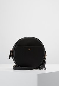 Kulatá černá kožená kabelka přes rameno se zlatými doplňky a střapcovým detailem. Hladká textura se zipem na zavření a nastavitelným popruhem.