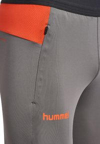 Close-up van grijze sportbroek met oranje meshpanelen, een zwart ritszakje en het oranje "hummel" logo op de dij.