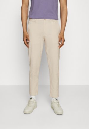 Personne portant un pantalon beige droit, des chaussettes blanches, des baskets blanches et une chemise violette rentrée, debout devant un fond blanc uni.