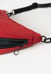 Borsa a tracolla rossa in tessuto con zip nera, tracolla regolabile e logo prominente. Presenta una superficie testurizzata e hardware resistente.