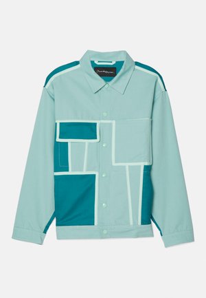Veste bleu clair dotée de patchs de différentes nuances de teal. Le design comprend des formes carrées et rectangulaires, avec des coutures et des boutons contrastants clairs.