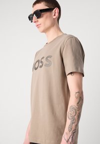 Šviesiai rudos spalvos marškiniai su apvalia iškirpte, juodo logotipo, trumpomis rankovėmis. Dėvintis turi juodus kvadratinius saulės akinius ir matomas rankų tetovėles.