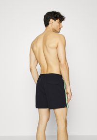 Pantaloni da nuoto neri con strisce laterali verdi, elastico in vita e tasca posteriore. Realizzati in tessuto leggero, presentano un design semplice.