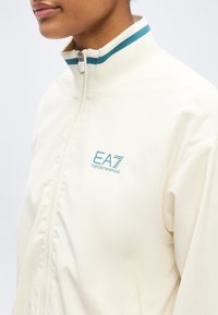 Λευκό μπουφάν με φερμουάρ και τιρκουάζ ρίγες στο γιακά, καθώς και τιρκουάζ λογότυπο "EA7 Emporio Armani" στο στήθος, που φοράει ένα άτομο.