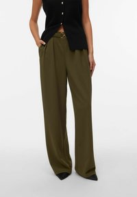 Vrouw draagt olijfgroene broek met wijde pijpen, voorplooien en een bijpassende riem, gecombineerd met een zwart mouwloos topje en zwarte puntige schoenen.