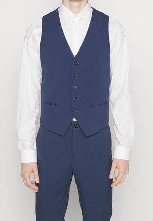 Homme portant un gilet boutonné bleu foncé et un pantalon assorti sur une chemise blanche, debout devant un fond uni.
