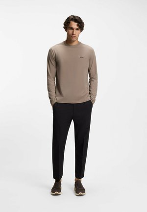 Long-sleeve beige top with a round neckline and small black logo, paired with black trousers and brown sneakers, set against a light background.