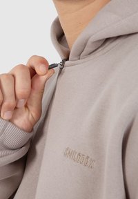 Lys beige zip-up hættejakke med ribbet kant og manchetter. Den har et lille broderet logo på venstre side og en nærbillede af lynlåsen i hånden.