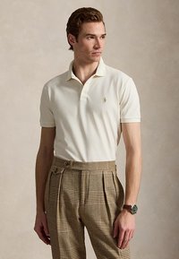 Homme portant un polo crème et un pantalon plissé marron à carreaux, regardant sur le côté avec une montre au poignet gauche sur un fond uni.