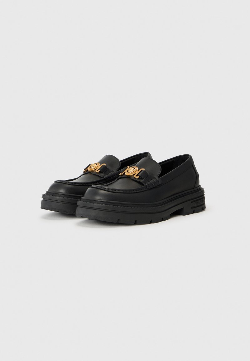 Mocassins en cuir noir avec une semelle épaisse, présentant un détail en émblème doré sur le devant et une texture lisse.