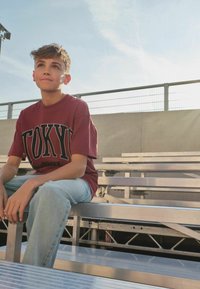 Teenager-Junge trägt ein bordeauxrotes Tokyo-T-Shirt und helle Jeans, sitzt auf einer Metalltribüne im Freien und schaut mit neutralem Gesichtsausdruck nach oben.