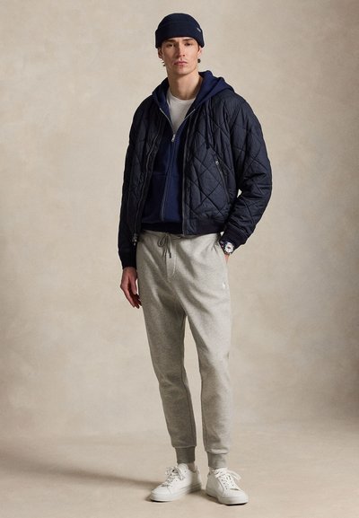 Jeune homme portant une veste matelassée bleu marine par-dessus un sweat à capuche bleu marine, un pantalon de jogging gris, des baskets blanches et un bonnet bleu marine, debout avec une main dans la poche.