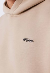 Sudadera con capucha beige hecha de tela suave con un bolsillo tipo canguro. Presenta un logo plateado brillante en el pecho izquierdo. Textura lisa, ajuste relajado.