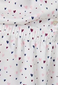 Tela blanca con cintura elástica fruncida, decorada con pequeños patrones de corazones en rosa, azul marino y burdeos, y un botón blanco visible.
