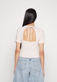 Top rosa claro com texturas, apresentando uma abertura nas costas com fecho de laço e mangas curtas, emparelhado com jeans azuis claros de perna larga.