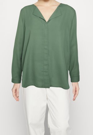 Blouse verte à manches longues avec un large col en V, associée à un pantalon blanc ample porté par une personne debout.