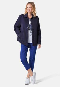 Marinejacke mit Knöpfen, zwei Seitentaschen, über einem weißen Hemd mit blauem Blumenmuster getragen; kombiniert mit blauen Cropped-Hosen und weißen Sneakern.