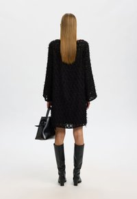 Svart texturerad klänning med långa ärmar, knälång, kombinerad med knähöga svarta stövlar och en svart handväska med cirkulärt handtag.