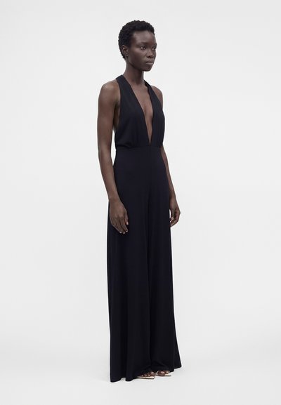 Personne mince à la peau foncée portant une combinaison noire sans manches à col halter avec jambes larges, se tenant devant un fond clair uni.