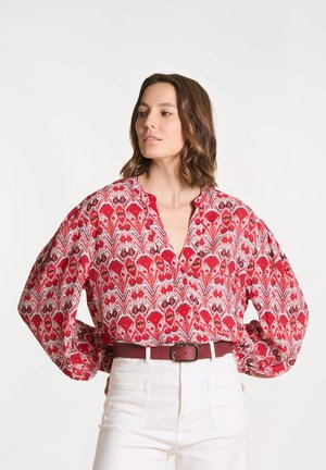 Femme portant un chemisier à manches longues rouge à motifs, rentré dans un pantalon blanc taille haute avec une ceinture bordeaux, debout devant un fond uni.