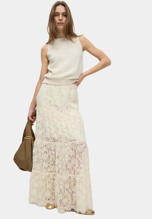 Donna che indossa un top senza maniche color crema e una lunga gonna di pizzo a strati, mentre tiene in mano una borsa marrone, in piedi davanti a uno sfondo chiaro e semplice.