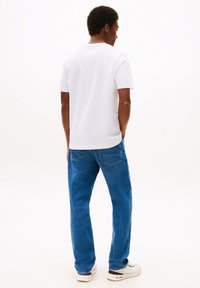 Witte katoenen T-shirt en blauwe denim jeans. De jeans zijn ruimvallend met twee achterzakken. Schoeisel zijn witte sneakers.