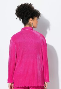 Giacca a maniche lunghe di colore rosa realizzata in tessuto a coste, con colletto, orlo ondulato e vestibilità morbida. Indossata sopra un completo coordinato.