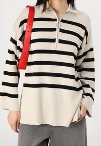 Suéter a rayas beige y negro con cuello con cremallera, ajuste oversized y mangas anchas; combinado con un bolso de cuero rojo de doble asa.