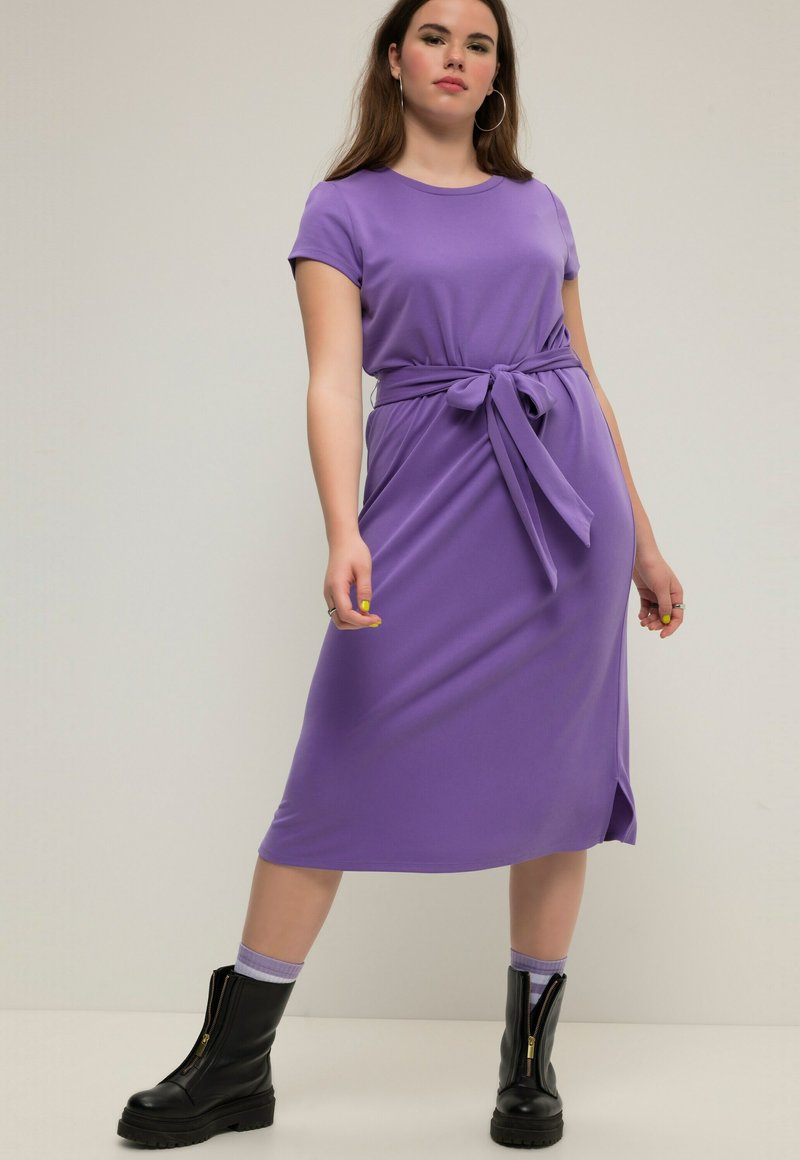 Lila Midi-Kleid mit kurzen Ärmeln und tailliertem Gürtel, glatter Textur und seitlichem Schlitz. Kombiniert mit schwarzen Stiefeletten und lila Socken.