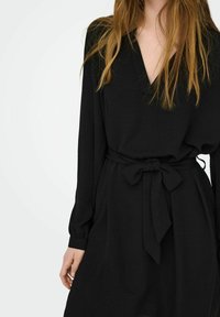 Black dress with long sleeves and a deep V-neck. Features a fabric belt tied at the waist. Smooth texture, simple, and minimalist design.