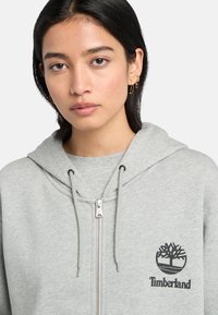 Grå lynlås-hættetrøje lavet af bomuldsblanding, med snor i hætten og et rundt Timberland-logo på brystet. Glat tekstur.