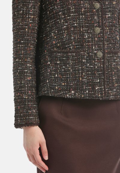 Rusin tummanruskea tweed-takki, jossa on monivärisiä pilkkuja. Varustettu kolmella napilla, kahdella etutaskulla ja aaltoilevilla hihoilla. Yhdistetty ruskeaan hameeseen.