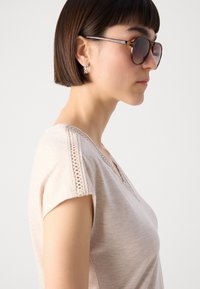 Beige top met korte mouwen en V-hals, voorzien van kantdetails op de schouders; gecombineerd met oversized schildpadbril en oorbellen.