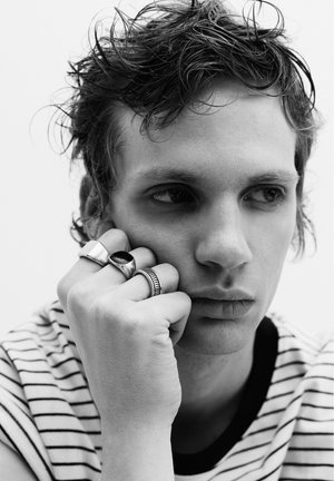 Jonge man met warrig haar, gekleed in een gestreept overhemd, rust zijn kin op een hand die versierd is met drie zilveren ringen, en kijkt peinzend opzij.