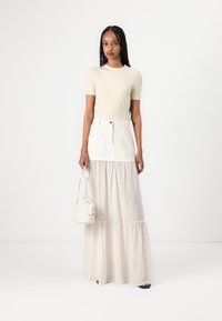 Top corto a maniche corte a coste color crema abbinato a una gonna maxi bianca a strati con tasche. Una piccola borsa leggera completa l'outfit.
