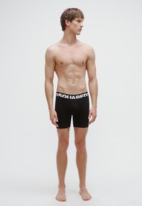 Črne bokserice z belim logotipom "DSQUARED2" na elastiki, z oprijetim krojem in elastičnim materialom, ki jih nosi stoječi moški model.