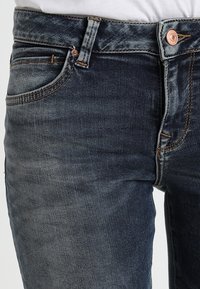 Tmavě modré džíny s bledým vzhledem, s měděnými knoflíky, pěti kapsami a kontrastním šitím podél švů.