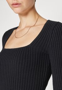 Kvinna som bär en svart ribbad tröja med fyrkantig halsringning och ett tunt guldhalsband, vilket visar halsen och  övre delen av bröstet.