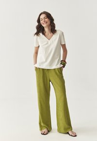 Fehér v-nyakú póló, amelyhez laza, textúrált, világoszöld, bő szabású nadrágot viselnek a modellen. A modell fekete szandált és színes karkötőket hord.