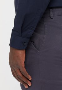 Camicia blu navy con bottoni e polsini abbottonati, abbinata a pantaloni grigi scuri con texture. In primo piano, una mano appoggiata sui pantaloni.